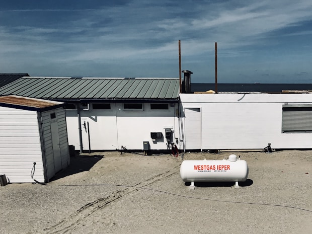 A beachside structure with a white exterior stands on a sandy area. A propane tank labeled 'WESTGAS IEPER' is positioned in front of the building. The roof is metal, and a clear blue sky and distant ocean are visible in the background.