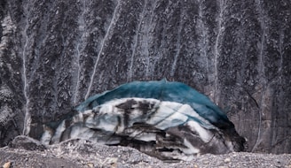 A glacier with layers of blue ice and dark, rocky sediment sits at the base of a steep rock face. The rock wall is streaked with vertical lines of erosion or mineral deposits, adding texture to the scene. The foreground shows a mix of loose rock and ice debris.