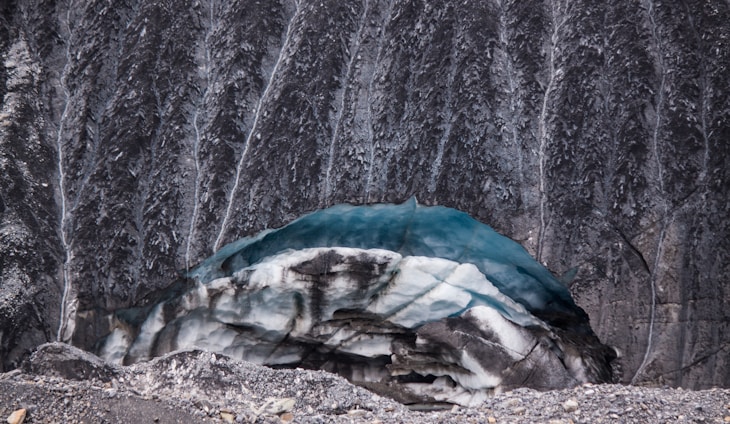 A glacier with layers of blue ice and dark, rocky sediment sits at the base of a steep rock face. The rock wall is streaked with vertical lines of erosion or mineral deposits, adding texture to the scene. The foreground shows a mix of loose rock and ice debris.