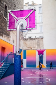 A vibrant urban basketball court set between tall buildings, featuring a pink and purple color scheme. The court is wet and reflective, indicating recent rain. Several people are present, engaging in a casual game of basketball.