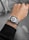A person is wearing a smartwatch on their left wrist. The watch has a silver mesh band and a digital display showing various circular icons and information, including temperature and calendar events. The person appears to be wearing a dark sweater.