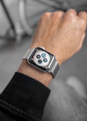 A person is wearing a smartwatch on their left wrist. The watch has a silver mesh band and a digital display showing various circular icons and information, including temperature and calendar events. The person appears to be wearing a dark sweater.