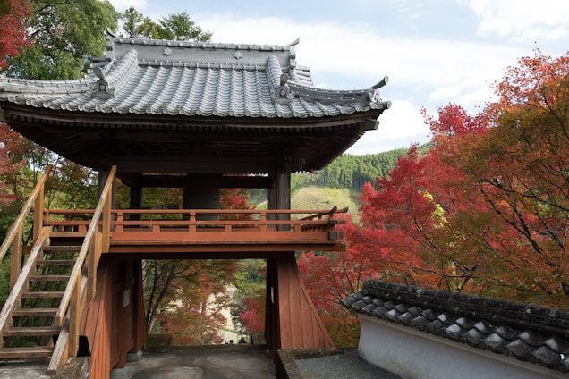 A traditional Japanese structure with a wooden staircase and intricately designed tiled roof is set among vibrant autumn foliage. The leaves display a range of colors from red to green, surrounding the building with a picturesque natural backdrop. The sky is partly cloudy, adding a serene atmosphere to the scene.