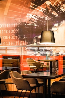 A modern interior space with contemporary furniture, featuring several chairs and a table. The background includes a wall mural with binary code and abstract shapes in warm tones. A black pendant light hangs from the ceiling, illuminating the tabletop area, and there are reflections and a menu on the table, adding to the ambiance.