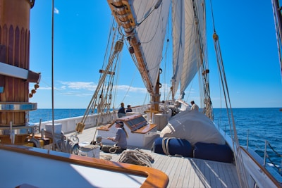 A view from the deck of a sailing ship with multiple sails unfurled, showing several crew members engaged in sailing activities. The clear blue sky and calm ocean stretch out to the horizon, providing a serene backdrop. Wooden elements and rigging dominate the foreground, contributing to the classic nautical atmosphere.