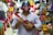 A person wearing a detailed helmet holds a small, colorful toy guitar. They stand in a vibrant market filled with various textiles and handmade crafts.