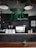 A modern coffee shop interior with a minimalist design. Hanging plants adorn the space, contrasting with dark, industrial elements such as a black corrugated metal wall and large, matte light fixtures. A coffee counter displays a shiny espresso machine, several menu cards, and neatly arranged utensils. A small potted plant sits on the counter, adding a touch of greenery.