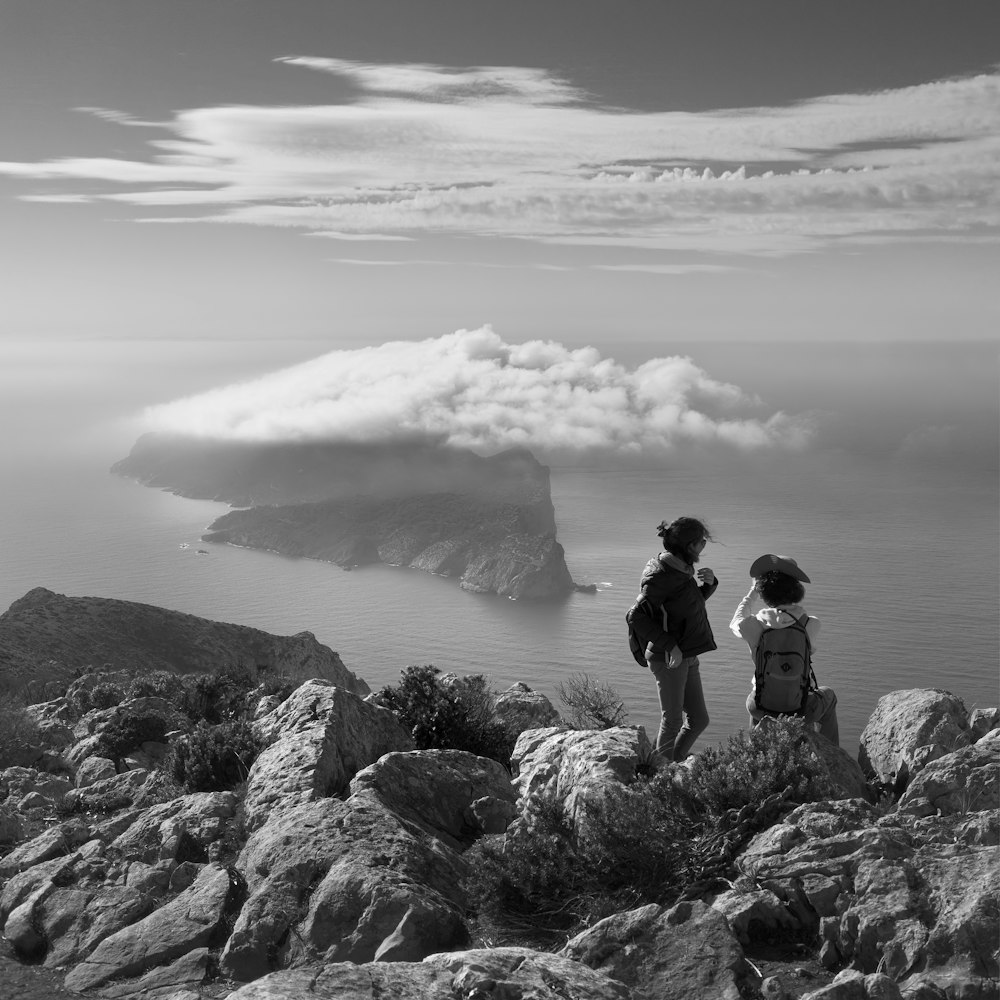 grayscale photo of two women on rock beside sea