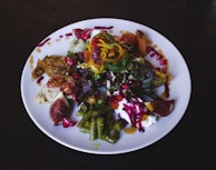 A colorful and vibrant salad arranged on a white plate, featuring a mix of ingredients such as figs, green beans, sliced peppers, pomegranate seeds, mixed greens, and dollops of cream. The dish is garnished with various sauces drizzled artistically around the plate, enhancing the presentation.