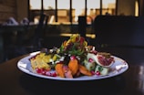 A gourmet plated dish features a variety of vibrant ingredients such as sliced figs, pomegranate seeds, colorful spiralized vegetables, and seasoned roasted carrots. The dish is presented on a white plate with an elegant, artistic arrangement, set against a dimly lit background with blurred restaurant interior.