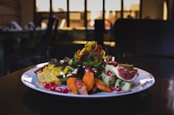 A gourmet plated dish features a variety of vibrant ingredients such as sliced figs, pomegranate seeds, colorful spiralized vegetables, and seasoned roasted carrots. The dish is presented on a white plate with an elegant, artistic arrangement, set against a dimly lit background with blurred restaurant interior.