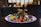 A gourmet plated dish features a variety of vibrant ingredients such as sliced figs, pomegranate seeds, colorful spiralized vegetables, and seasoned roasted carrots. The dish is presented on a white plate with an elegant, artistic arrangement, set against a dimly lit background with blurred restaurant interior.