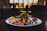 A gourmet plated dish features a variety of vibrant ingredients such as sliced figs, pomegranate seeds, colorful spiralized vegetables, and seasoned roasted carrots. The dish is presented on a white plate with an elegant, artistic arrangement, set against a dimly lit background with blurred restaurant interior.