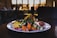 A gourmet plated dish features a variety of vibrant ingredients such as sliced figs, pomegranate seeds, colorful spiralized vegetables, and seasoned roasted carrots. The dish is presented on a white plate with an elegant, artistic arrangement, set against a dimly lit background with blurred restaurant interior.