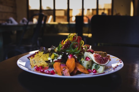 A gourmet plated dish features a variety of vibrant ingredients such as sliced figs, pomegranate seeds, colorful spiralized vegetables, and seasoned roasted carrots. The dish is presented on a white plate with an elegant, artistic arrangement, set against a dimly lit background with blurred restaurant interior.