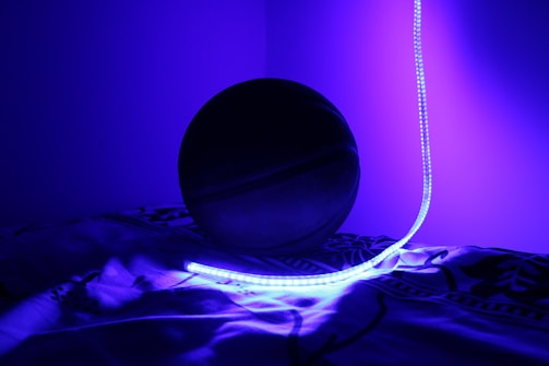 A basketball is partially illuminated by a bright, glowing blue LED light strip. The setting appears to be indoors, with the basketball resting on patterned fabric, enveloped in a deep blue hue.