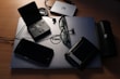 An assortment of electronic devices and personal items on a desk. A Dell laptop serves as a base, with a pair of glasses, a wallet, a closed PlayStation Portable, and a smartphone resting on it. Nearby, there is a JBL portable speaker, an external hard drive, and a Canon camera lens cap.