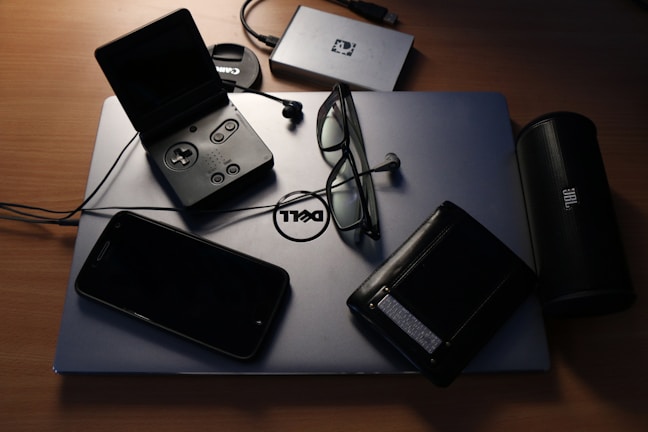 An assortment of electronic devices and personal items on a desk. A Dell laptop serves as a base, with a pair of glasses, a wallet, a closed PlayStation Portable, and a smartphone resting on it. Nearby, there is a JBL portable speaker, an external hard drive, and a Canon camera lens cap.