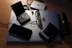 An assortment of electronic devices and personal items on a desk. A Dell laptop serves as a base, with a pair of glasses, a wallet, a closed PlayStation Portable, and a smartphone resting on it. Nearby, there is a JBL portable speaker, an external hard drive, and a Canon camera lens cap.