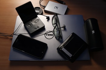 An assortment of electronic devices and personal items on a desk. A Dell laptop serves as a base, with a pair of glasses, a wallet, a closed PlayStation Portable, and a smartphone resting on it. Nearby, there is a JBL portable speaker, an external hard drive, and a Canon camera lens cap.