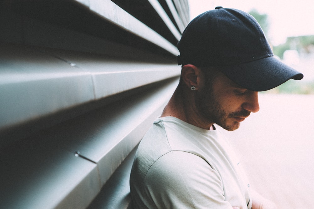 man wearing black cap and white shirt leaning on gray metal wall