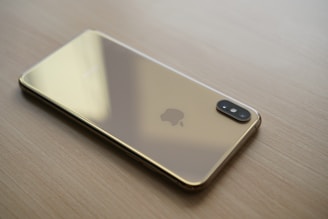 A sleek, gold-colored smartphone lies face down on a light wooden surface. The phone's back features a prominent logo and a dual camera setup aligned vertically in the top corner.