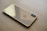 A sleek, gold-colored smartphone lies face down on a light wooden surface. The phone's back features a prominent logo and a dual camera setup aligned vertically in the top corner.