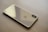 A sleek, gold-colored smartphone lies face down on a light wooden surface. The phone's back features a prominent logo and a dual camera setup aligned vertically in the top corner.