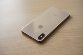A smartphone with a gold finish is placed face down on a light wooden surface, displaying its camera and Apple logo.