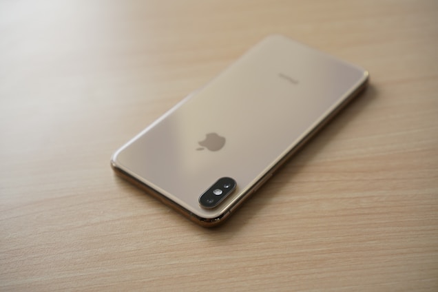 A smartphone with a gold finish is placed face down on a light wooden surface, displaying its camera and Apple logo.