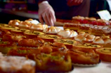 A variety of pastries are displayed on a table, including tarts, eclairs, and pies with different toppings like cream and nuts. The baked goods are arranged in rows, creating an appealing and colorful assortment. A person's hand wearing gloves is visible, possibly arranging or serving the pastries.