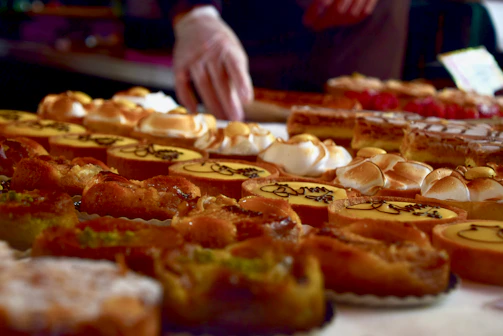 A variety of pastries are displayed on a table, including tarts, eclairs, and pies with different toppings like cream and nuts. The baked goods are arranged in rows, creating an appealing and colorful assortment. A person's hand wearing gloves is visible, possibly arranging or serving the pastries.