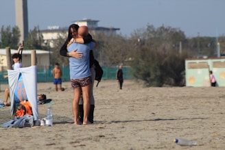 A beach setting where a man and woman embrace warmly in the foreground. Nearby, a towel or banner is draped over a wooden structure, next to a backpack and water bottles. Other people are dispersed in the background, some walking, others lounging. The atmosphere is relaxed with sparse greenery and buildings visible at a distance.