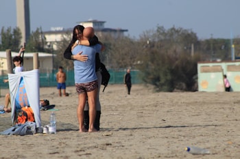 A beach setting where a man and woman embrace warmly in the foreground. Nearby, a towel or banner is draped over a wooden structure, next to a backpack and water bottles. Other people are dispersed in the background, some walking, others lounging. The atmosphere is relaxed with sparse greenery and buildings visible at a distance.