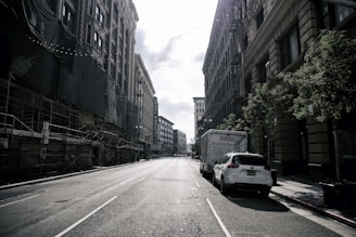 A deserted urban street flanked by tall buildings on both sides, with scaffolding visible on the left and parked vehicles, including a U-Haul truck, on the right. Strings of lights are suspended above the street, casting gentle shadows under the bright, partly cloudy sky. The buildings feature classic architectural details, and a few trees line the sidewalk.