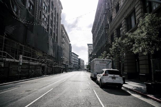 A deserted urban street flanked by tall buildings on both sides, with scaffolding visible on the left and parked vehicles, including a U-Haul truck, on the right. Strings of lights are suspended above the street, casting gentle shadows under the bright, partly cloudy sky. The buildings feature classic architectural details, and a few trees line the sidewalk.