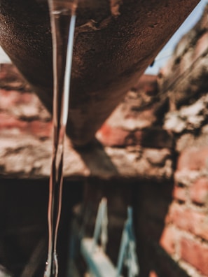 A narrow stream of water flows from a rusty pipe, with a background of red and brown bricks that are out of focus.