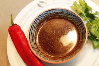 A patterned bowl filled with a rich, dark liquid sits on a plate. Beside it lies a vibrant red chili pepper, and a small bunch of fresh green cilantro leaves is placed nearby. The combination suggests ingredients for a spicy dish.