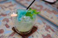 A glass filled with crushed ice and a blue popsicle, garnished with fresh mint leaves. The popsicle stick protrudes from the ice, and a black straw is also inserted into the glass. The surface on which the glass is placed has a weathered, rustic appearance with blue and brown hues.