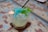 A glass filled with crushed ice and a blue popsicle, garnished with fresh mint leaves. The popsicle stick protrudes from the ice, and a black straw is also inserted into the glass. The surface on which the glass is placed has a weathered, rustic appearance with blue and brown hues.