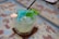 A glass filled with crushed ice and a blue popsicle, garnished with fresh mint leaves. The popsicle stick protrudes from the ice, and a black straw is also inserted into the glass. The surface on which the glass is placed has a weathered, rustic appearance with blue and brown hues.
