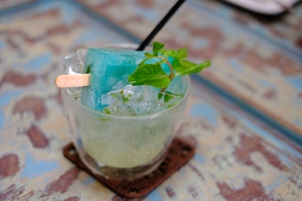 A glass filled with crushed ice and a blue popsicle, garnished with fresh mint leaves. The popsicle stick protrudes from the ice, and a black straw is also inserted into the glass. The surface on which the glass is placed has a weathered, rustic appearance with blue and brown hues.
