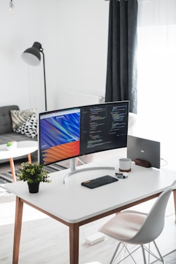 A modern home office setup with a sleek curved monitor displaying code and a colorful abstract background. A minimalist white desk holds a small black keyboard, a pair of wireless earbuds, a coffee mug, and a potted plant. Nearby, a sleek gray chair complements the room’s contemporary style, with a standing lamp and sofa in the background.