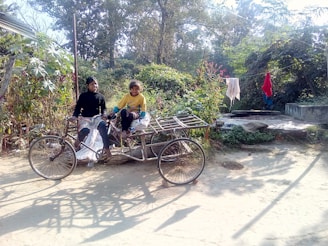 Two children are sitting on a cargo tricycle in a rural area. They are surrounded by lush greenery, and clothes are hanging to dry in the background. The scene is sunlit, with shadows cast on the sandy path.