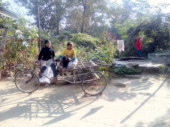 Two children are sitting on a cargo tricycle in a rural area. They are surrounded by lush greenery, and clothes are hanging to dry in the background. The scene is sunlit, with shadows cast on the sandy path.