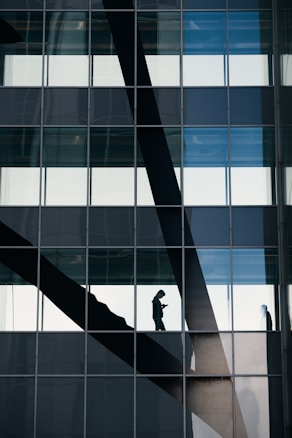 A grid of glass windows covers a building facade, reflecting the outside environment with a visible silhouette of a person walking across a staircase inside.