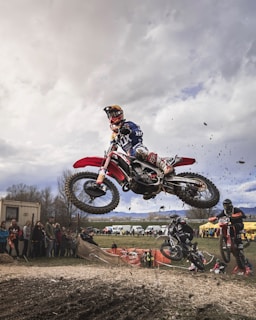 Action shot of a motocross rider mid-jump over a dirt track with spectators in the background.