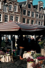 A busy street market scene with a striped canopy sheltering a variety of colorful flowers displayed in crates. The market is set against a backdrop of ornate, historic European-style buildings. Shadows cast by vendors and shoppers suggest a bustling atmosphere.