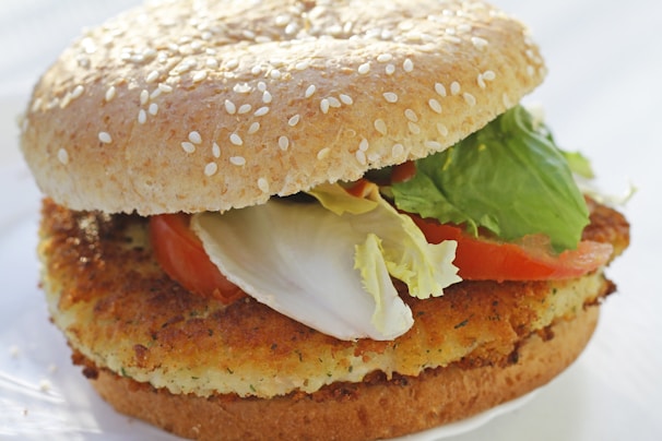 A close-up view of a sandwich featuring a sesame seed bun with a crispy patty, fresh lettuce, and slices of tomato. The ingredients are stacked neatly, highlighting a variety of textures and colors.