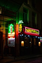 The image features a dimly lit street scene with neon signs of a Chinese restaurant called 'La Tour de Jade' in vibrant red, green, and yellow colors. The signs include both English and Chinese characters. The storefront is adorned with glowing lights highlighting the entrance and windows, creating a lively and inviting atmosphere.